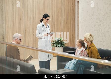 Kaukasische Ärztin mittleren Alters stehend und in der Hand eines digitalen Tablets, während sie mit einem kaukasischen älteren Mann und einer kaukasischen Frau mit Kind auf dem Sofa im Wartezimmer sprach Stockfoto