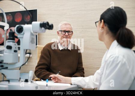 Porträt eines älteren kaukasischen Mannes, der in der Augenklinik sitzt und in die Kamera schaut, während junge Erwachsene asiatische Frau Doktor über die Ergebnisse der Augenuntersuchung mit medizinischen Geräten diskutiert Stockfoto