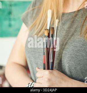 Junge kaukasische Frau, die Pinsel in einem Kunststudio hält und ein graues T-Shirt trägt Stockfoto