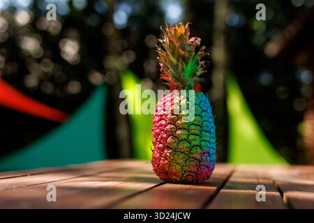 Hell bemalte Ananas auf einem Holztisch im Sonnenlicht, mit farbenfrohen Hintergrunddekorationen. Stockfoto