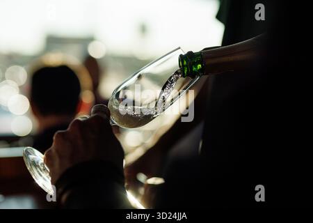 Nahaufnahme eines Champagners, der aus einer Flasche in ein Glas gegossen wird, mit Bokeh-Lichtern und Menschen in weichem Hintergrund. Stockfoto