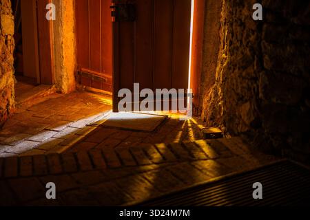 Warmes Licht, das durch eine teilweise geöffnete Holztür in einem alten Steininneren leuchtet. Stockfoto