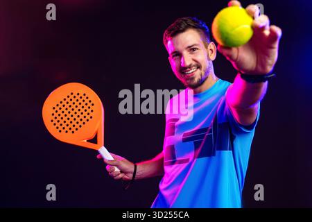 Junger Mann spielt Padel-Tennis mit Schläger und Ball in farbenfroher Beleuchtung Stockfoto