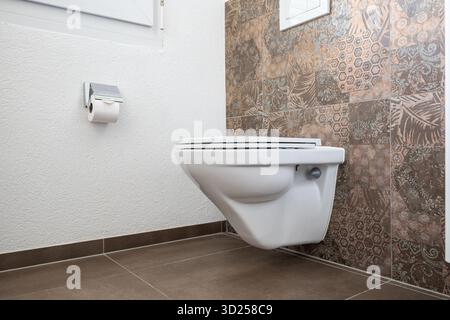 Weiße Keramik-Toilettenschüssel im modernen Bad mit gefliestem Boden und Wand. Seitenansicht, niedriger Winkel, keine Personen, geschlossener Deckel Stockfoto