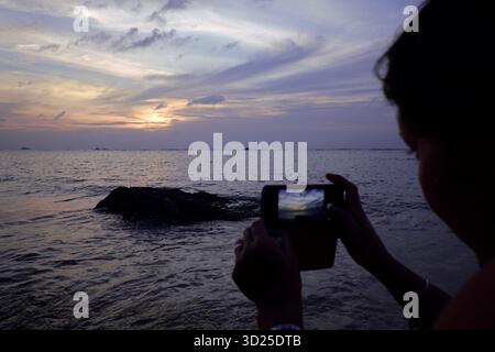 Kaukasische Frau fotografiert den Sonnenuntergang mit dem Smartphone, steht in der Nähe der Küste mit sichtbaren Wellen und Felsen und nimmt während der Dämmerung mit Wolken am Himmel eine malerische Aussicht auf Stockfoto