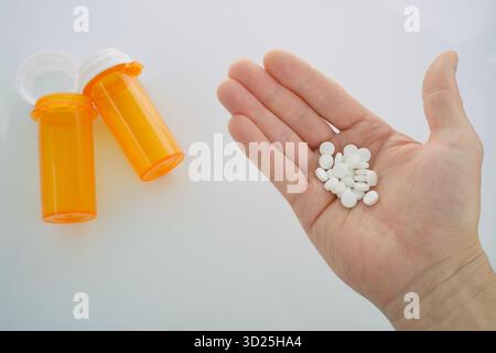 Ansicht von oben nach unten: Hand eines Mannes, der einen Stapel weißer, runder Tabletten hält, mit zwei leuchtend orangen verschreibungspflichtigen Flaschen im Hintergrund. Stockfoto