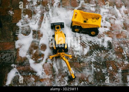 Zwei gelbe Spielzeugwagen ruhen auf einer verschneiten Ziegeloberfläche und zeigen Winterspiele und Kreativität an einem kalten Tag. Stockfoto