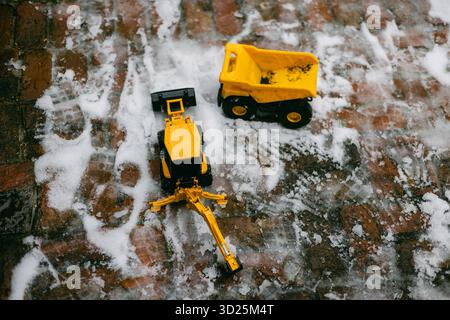Gelbe Spielzeugwagen sitzen auf einem gemauerten Gehweg im Schnee und zeigen spielerischen Winterspaß in der kalten Jahreszeit. Stockfoto