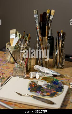 Malpinsel und Farbpalette des Künstlers auf einem unordentlichen Studiotisch mit Tuben aus Farbe und Skizzenbuch Stockfoto