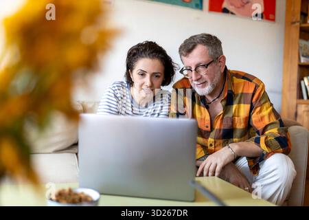 Ein Paar mittleren Alters sitzt eng zusammen auf einem Sofa und schaut aufmerksam auf einen Laptop-Bildschirm auf einem Couchtisch und deutet an, dass sie arbeiten, einkaufen oder arbeiten Stockfoto