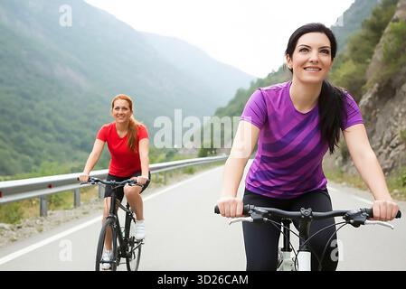 Zwei Frauen fahren eine Straße hinunter mit dem Fahrrad. Einer trägt ein rotes Hemd und der andere ein lila Hemd. Sie lächeln und genießen die Fahrt Stockfoto