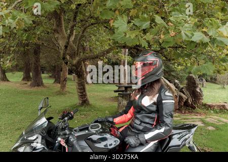 Motorradfahrerin mit Schutzausrüstung und Helm, die in einem grünen Park auf dem Motorrad sitzt Stockfoto