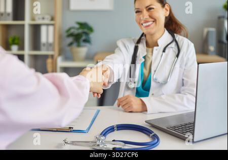 Glückliche lächelnde Ärztin sitzt am Schreibtisch und schüttelt den Patienten die Hand für freundliche Interaktion in der Klinik Stockfoto