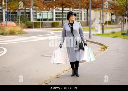 Eine Frau, die einen stilvollen grauen Mantel und Hut trägt, läuft im Herbst fröhlich auf einer Stadtstraße entlang. Sie trägt mehrere Einkaufstaschen und genießt ihren Tag im Freien Stockfoto