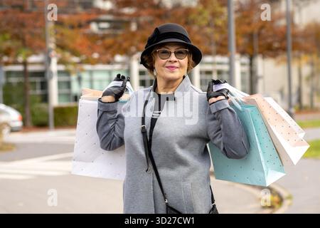 Eine stilvolle Frau in grauem Mantel und Hut trägt mehrere Einkaufstaschen, während sie im Herbst in einem Park spaziert. Die bunten Taschen spiegeln ihr fröhliches Moo wider Stockfoto