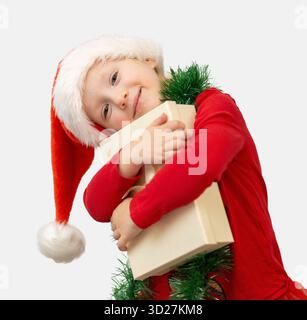 Ein fröhliches Kind in rotem Hemd und Weihnachtsmann-Hut umhüllt ein Geschenk, das während der Weihnachtszeit zu Hause Spannung und Glück ausstrahlt. Stockfoto