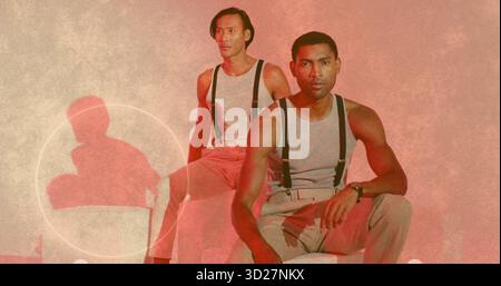 Zwei Männer mit grauen Oberteilen, beigefarbenen Hosen und Hosenträgern auf Studiohockern, die Armbanduhr zeigen Stockfoto