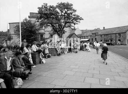 Ein Schwarzweißfoto fängt eine geschäftige Szene in einer kleinen englischen Stadt ein. Eine Gruppe von Fußgängern, vor allem Erwachsene und Kinder, ist auf einem gepflasterten Platz versammelt und wartet auf einen Bus. Die Szene wird von einem prächtigen Gebäude, „The Royal Oak“, und alten Bäumen dominiert, die einen Einblick in das tägliche Leben und den architektonischen Charme einer historischen Stadt in Großbritannien bieten. Stockfoto