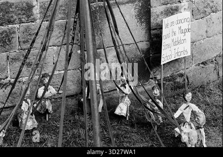 Dieses Schwarzweiß-Foto zeigt eine gut gekleidete Ausstellung in Chesterfield, Großbritannien. Das Bild zeigt eine Sammlung von Steinfiguren, die an Holzpfählen aufgehängt sind, die an einer Steinmauer angeordnet sind. Im Hintergrund ist ein Schild mit der Aufschrift „Wir tanzen um die Maypole“ zu sehen, das auf die jährliche Well Dressing-Veranstaltung hinweist. Stockfoto
