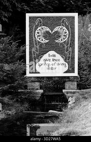 Dieses Bild zeigt ein Schild, das im Rahmen des Annual Well Dressing in Chesterfield, Großbritannien, errichtet wurde. Auf dem Schild steht „Ich gebe dir Quellen lebendigen Wassers“ und eine stilisierte Darstellung eines Wassertropfens. Die Szene wird in Schwarz-weiß präsentiert und zeigt die traditionelle künstlerische Darstellung, die mit der Veranstaltung verbunden ist. Stockfoto