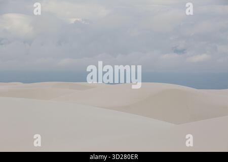 Weiche, glatte Sanddünen erstrecken sich über die Landschaft, während dunkle Wolken über sich ziehen. Die Szene fängt die Ruhe der Natur bei Sonnenaufgang in White Sands ein Stockfoto
