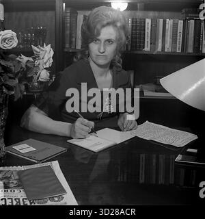 Archiv Schwarzweißfoto aus den 1970er Jahren in Sloviansk, Ukrainische SSR. Sie zeigt Deputy Chukhleb in ihrem Büro oder Arbeitszimmer. Sie sitzt an einem polierten Schreibtisch, blickt direkt in die Kamera, während sie einen Stift über ein offenes Notizbuch hält, als ob sie Notizen macht. Hinter ihr befindet sich ein großer Bücherregal voller Bücher. Links von ihr befindet sich eine Vase mit Rosen, die dem offiziellen Setting eine sanftere Note verleiht. Sie trägt ein formelles Kleid mit einem Deputy-Abzeichen. Dieses Porträt zeigt eine typische Sowjetfrau in einer Position der Autorität während der 1970er Jahre Stockfoto