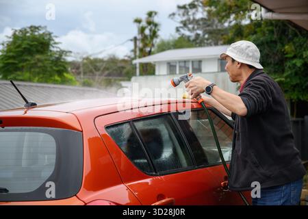 Mann, der das Auto auf der Auffahrt mit einer Bürste und einem Wasserspray aus einem Schlauch wäscht. Stockfoto