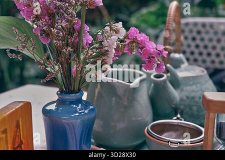 Stillleben aus Keramik-Teekannen und frischen rosa Blumen in einer blauen Vase, die im Freien bei weichem Tageslicht in einem gemütlichen Gartencafé festgehalten werden. Stockfoto