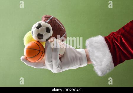 Weihnachtsmann Hand im Anzug und Handschuhe, die kleine Bälle verschiedener Sportarten auf grünem isoliertem Hintergrund halten. Weihnachtswettbewerbkonzept verschiedener Bälle Stockfoto