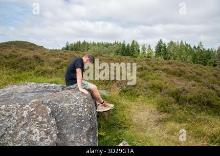 Einsamer Mann sitzt auf einem großen Felsen traurig, blickt auf die Füße in der Heidelandschaft ein Sommertag, Themenbild mit Kopierraum Stockfoto