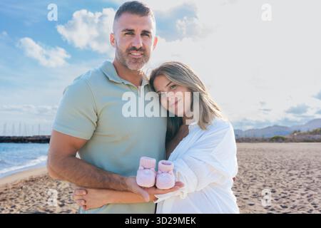 Glückliches Paar am Strand, schwangere Frau, die Babyschuhe hält, in die Kamera lächelt. Stockfoto