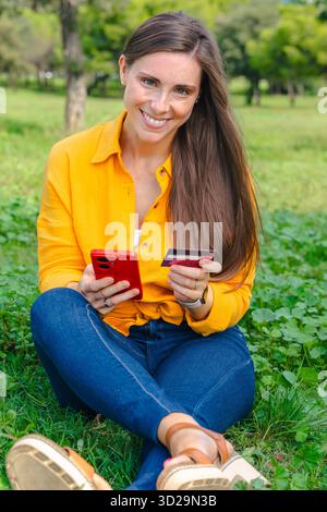 Lächelnde Frau draußen, die Smartphone und Kreditkarte hält. Stockfoto