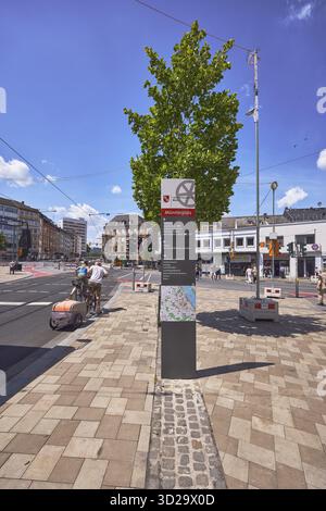Stele mit Ortsschild, Verkehrsgebiet, Straße, Platz, Bürgersteig aus Betonpflastersteinen, allgemeine Architektur, Radfahrer und Fußgänger als a s Stockfoto