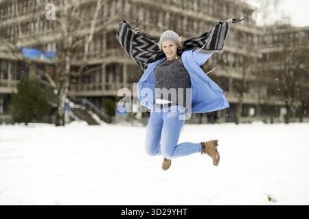 Frau in Winterkleidung, die bei neuem Wetter in die Luft springt, mit fröhlichem Lächeln, glücklich über den Schnee, den sie liebt, euphorischer Sprung, Arme ausgestreckt W Stockfoto