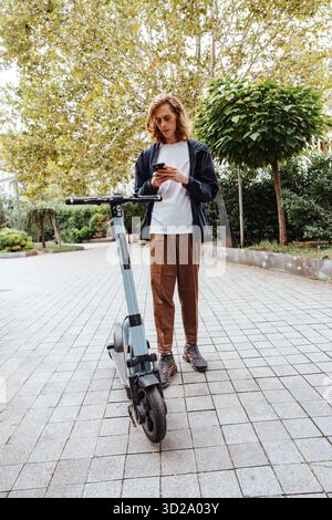 Junger Erwachsener, der im Stadtpark neben einem Elektroroller telefoniert Stockfoto