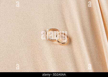 Zwei goldene Eheringe liegen auf einem goldenen glänzenden Hintergrund aus schickem Stoff mit weichen Falten von Vorhängen. Eine Kopie des Bereichs. Mockup für Hochzeitsdesign Stockfoto