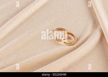 Zwei goldene Eheringe liegen auf einem schicken goldenen Stoffhintergrund mit sanften Wellen. Ein Kopierbereich. Mockup für Hochzeitsdesign. Einladung. Postkarte Stockfoto