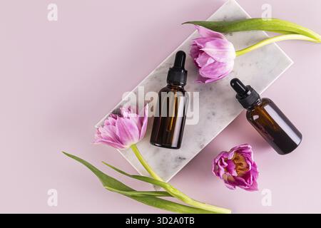 Zwei Kosmetikflaschen mit einer Pipette eines natürlichen Bio-Produkts liegen auf einem Podium aus weißem Marmor mit feinen Tulpen. Leeres Layout. Verpackung Stockfoto
