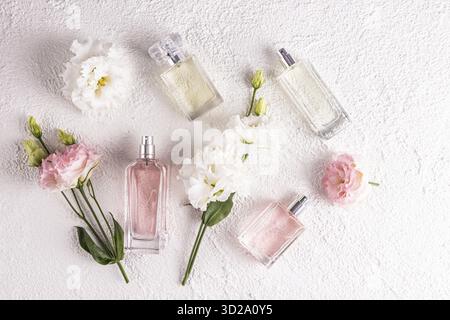 Mehrere schöne Parfümflaschen in verschiedenen Formen unter Blumen auf weißem Reliefhintergrund. Draufsicht. Flache Lagen. Präsentation von Duftstoffen Stockfoto