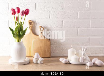 Osterhintergrund, Vorderansicht auf dem Tisch, dekoriert mit Blumen in einer Vase, traditionellen Eiern und Keramikkaninchen, Platz für einen Text. Weiße Ziegelwand Stockfoto