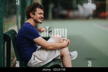 Entspannter Mann sitzt auf einer Parkbank in der Nähe eines Tennisplatzes und genießt einen ruhigen Moment im Freien Stockfoto