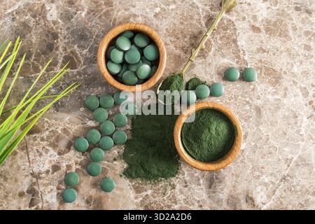 Grünes Spirulina-Pulver und Tabletten in Holzschalen auf braunem Marmorboden. Ein beliebtes Superfood für Vegetarier und Sportler. Draufsicht Stockfoto