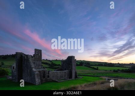 Idyllische irische Landschaft mit friedlichem Sonnenuntergang und historischen Ruinen der Fore Abbey. Ruhiger Blick in die Dämmerung mit antiken Abteiresten umgeben von sanften Sonnenuntergangsfarben und sanftem Himmel, Irland Stockfoto