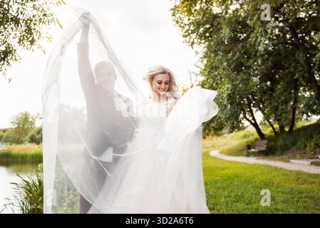 Glückliche Braut und Bräutigam am Hochzeitstag, langes Brautschleier heben, im Sommerpark spazieren gehen || Model veröffentlicht Stockfoto