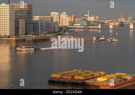 Ein Uber Boat Thames Clipper River Bus kommt am Royal Wharf Quay an, während ein anderer am Nordufer der Themse mit den Thames Barrier-Hochwasserschutztoren im Hintergrund in London, Großbritannien, am 15. April 2023 abfährt. Im Vordergrund befinden sich zwei schwimmende passive Treibholzsammler (PDCs), die Käfige sind, um Treibholz, Ablagerungen und Kunststoffabfälle aus dem Fluss zu sammeln. Quelle: Rob Taggart/Alamy Stockfoto