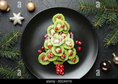 Weihnachtsbaum aus Kiwi, Granatapfelkernen und festlicher Dekoration auf schwarzem Tisch, flache Ladefläche Stockfoto