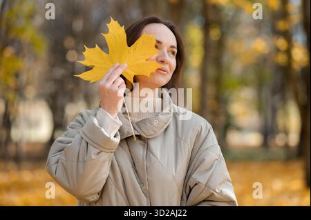 Die lächelnde Frau in einer beigefarbenen Jacke hält ein großes gelbes Ahornblatt nahe ihrem Gesicht, während sie in einem Park steht, der mit goldenem Herbstlaub gefüllt ist Stockfoto