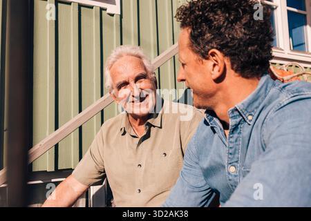 Senior Mann, der mit dem Sohn spricht, während er an sonnigen Tagen auf Stufen vor der Villa sitzt Stockfoto