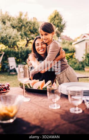 Lächelnde Mutter, die Tochter umschließt, während sie am Esstisch im Garten sitzt Stockfoto