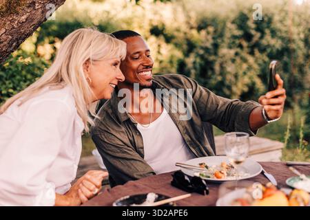 Glücklicher Mann, der Selfie mit Mutter macht, während er am Esstisch im Hinterhof sitzt Stockfoto